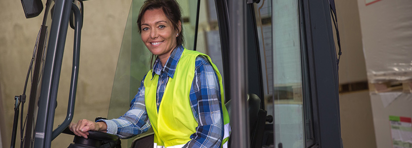 A photograph of girl with a yellow safety west sitting on the forklift.