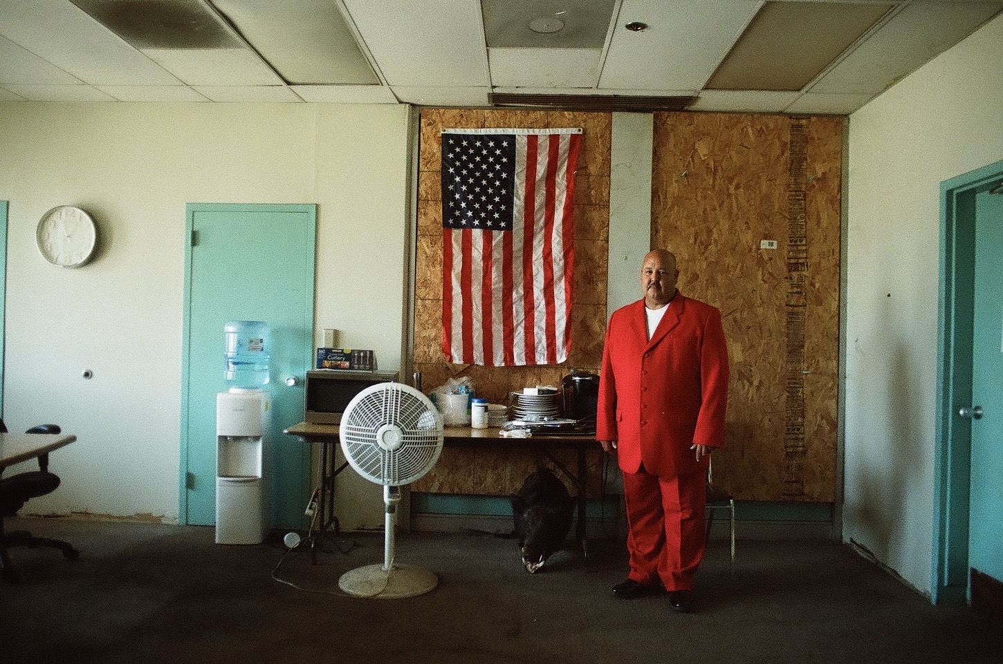 photograph showing a man wearing a red suit, standing in front of a wall. There is an american flag hanging behind him.