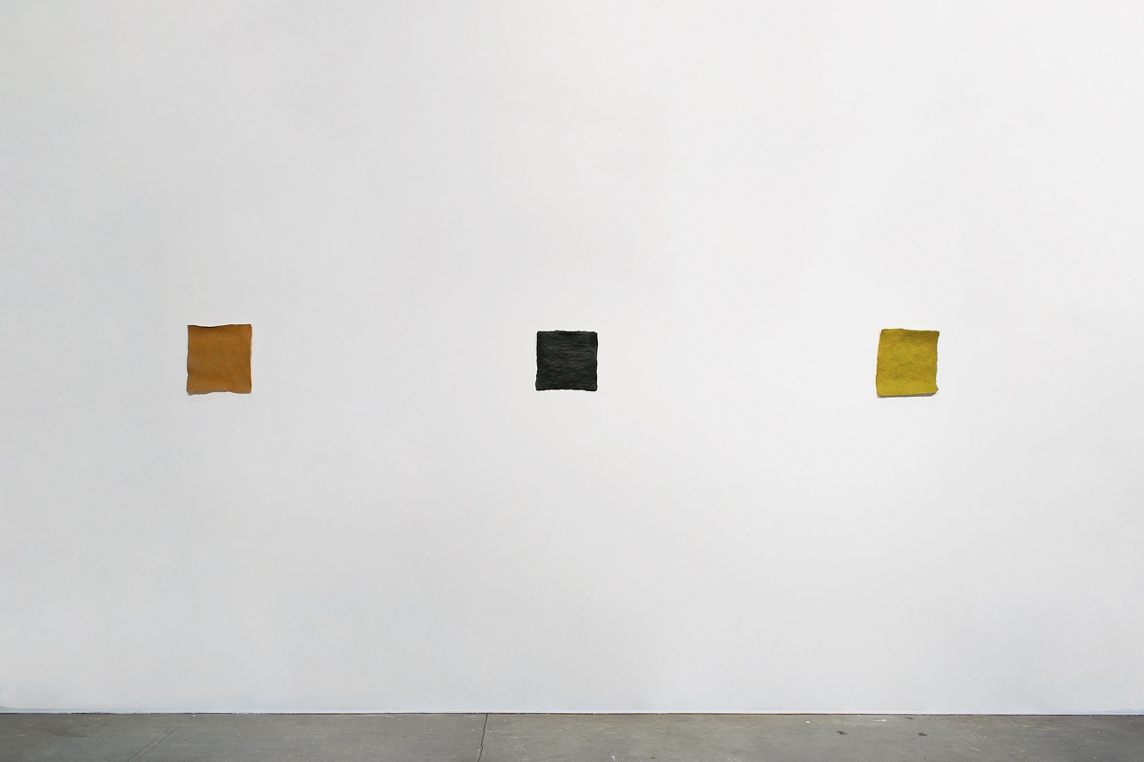 installation view of three irregular square shaped artworks in tones of ochre, black, and red against a white wall. 
